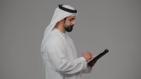 إنجاز العمل عن بعد عبر جاز التابليت المحمول، ايماءات التركيز والاندماج , استخدام التكنولوجيا الحديثة ،بورتريه لرجل عربي خليجي اماراتية ترتدي الكندورة والغترة يقوم بالكتابة وتدوين الملاحظات على الجهاز اللوحي ،دمج التقنيات الحديثة في الحياة العملية،  خلفية رمادية