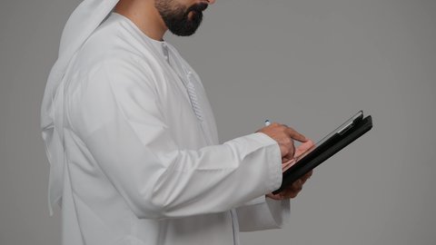 Working remotely via a portable tablet, gestures of concentration and integration, using modern technology, portrait of an Emirati Gulf Arab man wearing a kandura and ghutra writing and taking notes on a tablet, integrating modern technologies into practical life, gray background