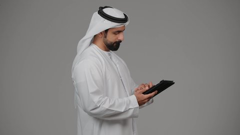 عقد اجتماع عمل عن بعد عبر الجهاز اللوحي، التواصل مع الاهل والاصدقاء، دمج التقنيات والتكنولوجيا الحديثة بالحياة اليومية، بورتريه من الجانب لرجل عربي خليجي اماراتي يرتدي الكندورة و الغترة يقوم بإجراء مكالمة فيديو عبر التابليت المحمول، خلفية رمادية.