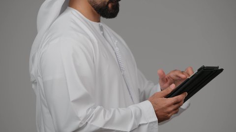 Browsing social media on a tablet, communicating with family and friends, integrating modern technology into daily life, close-up side portrait of an Emirati Gulf Arab man wearing a kandura and ghutra using a tablet, gray background.