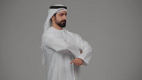 مفهوم الاناقة والرجولة والوسامة، الاهتمام بالمظهر الخارجي، بورتريه لرجل عربي خليجي إماراتي مبتسم يرتدي الكندورة الاماراتية والغترة يقف بأيدي متقاطعة وبنظر الى الكاميرا بإيماءات السرور،  خلفية رمادية