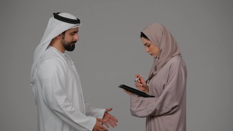 ازمة في العلاقات الاسرية، رجل إماراتي يقوم بتوبيخ زوجته بإيماءات الغضب، مشاكل عائلية وصراع اسري، شجار بين الزوجين، امرأة محجبة ترتدي العباءة تقوم بالكتابة على الجهاز اللوحي المحمول، بورتريه لزوجين عربيين خليجيين اماراتيين يتجادلان مع بعضهما، خلفية بيضاء