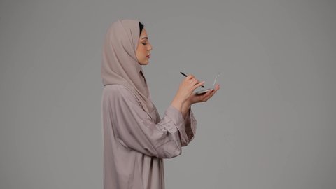 مفهوم الانوثه والاهتمام بالمظهر الخارجي، سيدة اماراتية تستخدم أدوات تجميل نسائيه عصريه، مكياج وميكاب تتوريال، التسويق لمنتجات التجميل، بورتريه من الجانب لامرأة عربيه سعودية خليجيه ترتدي العباءة والحجاب تحمل بيدها فرشاة توريد الخدود، خلفية بيضاء