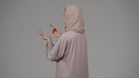 مفهوم الانوثه والاهتمام بالمظهر الخارجي، سيدة اماراتية تستخدم أدوات تجميل نسائيه عصريه، مكياج وميكاب تتوريال، التسويق لمنتجات التجميل، بورتريه من الجانب لامرأة عربيه سعودية خليجيه ترتدي العباءة والحجاب تحمل بيدها فرشاة توريد الخدود، خلفية بيضاء