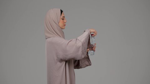 المحافظة على سلامة الجهاز المناعي للجسم , المداومة على شرب المياه ، بورتريه من الجانب لامراة عربية خليجية امارتية محجبة ترتدي العباءة تقوم بالشرب من زجاجة ماء بلاستيكية صغيرة, سيدة سعودية تشعر بالعطش والجفاف , خلفية رمادية