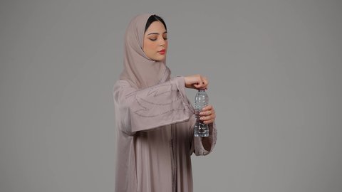 المداومة على شرب المياه ، المحافظة على سلامة الجهاز المناعي للجسم , بورتريه لامراة عربية خليجية امارتية محجبة ترتدي العباءة تقوم بالشرب من زجاجة ماء بلاستيكية صغيرة, سيدة سعودية تشعر بالعطش والجفاف , خلفية رمادية