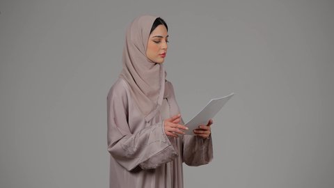 Writing down daily tasks, reading notes and information, following up on plans and projects, work papers, portrait of a veiled Arab Gulf Emirati woman wearing an abaya holding white papers in her hands, a young Saudi woman doing scientific research, gestures of seriousness and self-confidence, gray background