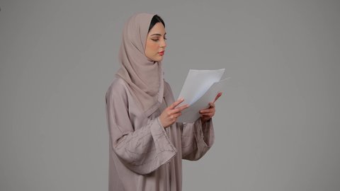 قراءة الملاحظات والمعلومات، متابعة الخطط والمشاريع ، أوراق خاصة بالعمل, تدوين المهام اليومية، بورتريه لامراة عربيه خليجيه اماراتيه محجبه ترتدي العباءة تحمل بيديها أوراق بيضاء، شابه سعوديه تقوم بالبحث العلمي، ايماءات الجدية والثقة بالنفس، خلفية رمادية
