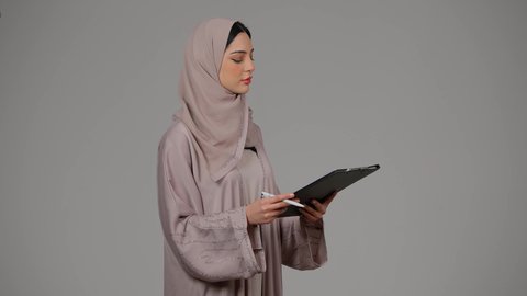 ايماءات الجدية والثقة بالنفس، تدوين المهام اليومية، قراءة الملاحظات والمعلومات، متابعة الخطط والمشاريع ، أوراق خاصة بالعمل, بورتريه لامراة عربيه خليجيه اماراتيه محجبه ترتدي العباءة تحمل بيديها أوراق بيضاء، شابه سعوديه تقوم بالبحث العلمي، خلفية رمادية