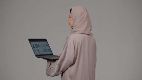 تصفح الإنترنت وإستخدام اجهزة تقنية حديثة، إدارة شؤون الأعمال، بورتريه من الجانب لشابة عربية خليجية اماراتية تقوم بالعمل على الحاسوب المحمول، العمل لتحقيق الاهداف، سيده سعودية تستخدم الحاسوب المحمول لإنجاز المهام، خلفية رمادية
