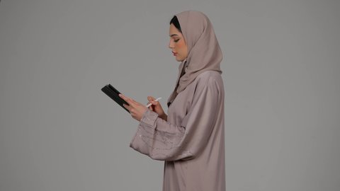 Integrating modern technologies into practical life, expressions of integration and focus, side portrait of a veiled Saudi Arabian Gulf woman wearing an abaya using a tablet pen, an Emirati woman recording and taking notes on a tablet, completing work remotely, gray background