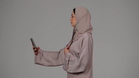 الاناقة والاهتمام بالمظهر الخارجي، التكنولوجيا والتقنية الحديثة والمتطورة، بورتريه لامرأة عربية خليجية اماراتية محجبة ترتدي العباءة تقوم باستخدام الهاتف المحمول، سيدة اماراتية بالتقاط صورة سيلفي تذكارية, خلفية رمادية