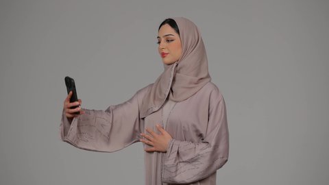 الاناقة والاهتمام بالمظهر الخارجي، التكنولوجيا والتقنية الحديثة والمتطورة، بورتريه لامرأة عربية خليجية اماراتية محجبة ترتدي العباءة تقوم باستخدام الهاتف المحمول، سيدة اماراتية بالتقاط صورة سيلفي تذكارية, خلفية رمادية
