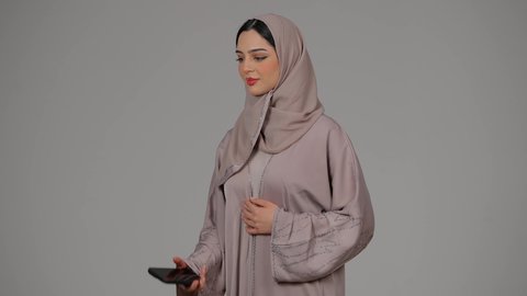 Communication and reassurance about family and friends, modern and advanced technology, portrait of a veiled Emirati Gulf Arab woman wearing an abaya using a mobile phone, an Emirati woman making a phone call on a mobile phone with expressions of happiness and pleasure, gray background