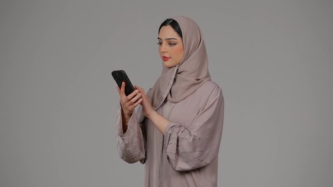 Modern and advanced technology, communication and reassurance about family and friends, portrait of a veiled Emirati Gulf Arab woman wearing an abaya using a mobile phone, an Emirati woman making a phone call on a mobile phone with expressions of happiness and pleasure, gray background