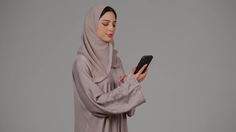 Communication and reassurance about family and friends, modern and advanced technology, portrait of a veiled Emirati Gulf Arab woman wearing an abaya using a mobile phone, an Emirati woman making a phone call on a mobile phone with expressions of happiness and pleasure, gray background
