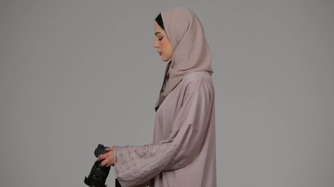 احتراف فن التصوير الفوتوغرافي باستخدام كاميرات رقمية عالية الدقة, وظيفة ومهنة نسائية, بورتريه من الجانب لإمراة عربية خليجية اماراتية ترتدي العباءة تقوم بالتقاط صورة تذكارية عبر كاميرا فوتوغرافية, مصورة سعودية تمارس هواية التصوير بتعابير السرور، خلفية رمادية