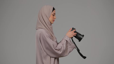 احتراف فن التصوير الفوتوغرافي باستخدام كاميرات رقمية عالية الدقة, وظيفة ومهنة نسائية, بورتريه من الجانب لإمراة عربية خليجية اماراتية ترتدي العباءة تقوم بالتقاط صورة تذكارية عبر كاميرا فوتوغرافية, مصورة سعودية تمارس هواية التصوير بتعابير السرور، خلفية رمادية