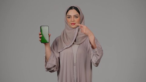 الاشارة بإصبع السبابة إلى شاشة الجوال، سيدة اماراتية تعرض شاشة خضراء فارغة، كروما ، استخدام التكنولوجيا والتقنية الحديثة،  تصفح مواقع التواصل الاجتماعي، بورتريه لامرأة عربية خليجية اماراتية محجبة ترتدي العباءة تقوم بإستخدام الهاتف المحمول، خلفية بيضاء