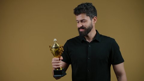 تشجيع الأنديه والمنتخبات، أنشطة ترفيهية ممتعة، النظر إلى الكاميرا بإيماءات الفرح والسعادة لفوز الفريق المفضل، رجل عربي خليجي اماراتي مبتسم يرتدي الزي الكاجوال يقفز عاليا ويمسك بيده الكأس الذهبي، تعابير الانتصار والحماس، خلفية صفراء