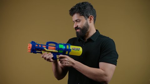 تعابير الحماس والفرح، مفهوم الترفيه عن النفس والتسلية،  قضاء وقت ممتع بالألعاب المائية في فصل الصيف، تصوير لشاب عربي خليجي اماراتي يرتدي الزي الكاجوال يحمل مسدس مائي بلاستيكي يقوم باللعب بإيماءات السرور والسعادة، خلفية ضفراء