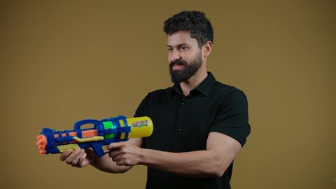 قضاء وقت ممتع بالألعاب المائية في فصل الصيف، تصوير لشاب عربي خليجي اماراتي يرتدي الزي الكاجوال يحمل مسدس مائي بلاستيكي يقوم باللعب بإيماءات السرور والسعادة، تعابير الحماس والفرح، مفهوم الترفيه عن النفس والتسلية، خلفية ضفراء