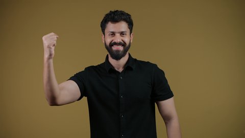 أنشطة ترفيهية ممتعة، النظر إلى الكاميرا بإيماءات الفرح والسعادة لفوز الفريق المفضل، تشجيع الأنديه والمنتخبات، رجل عربي خليجي اماراتي مبتسم يرتدي الزي الكاجوال يرفع قبضة اليد بتعابير الانتصار والحماس، خلفية صفراء