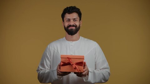 مفهوم الشكر والامتنان، التعبير عن الحب والتقدير، تقديم الهدايا بالاعياد والمناسبات الخاصة، النظر إلى الكاميرا بايماءات السرور، تصوير لشاب عربي خليجي اماراتي يرتدي الكندورة يحمل بيديه صندوق هدية، خلفية صفراء