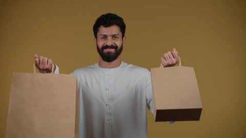 Taking advantage of discounts and clearance sales, commercial purchases and gifts, looking at the camera with happy expressions, shopping and taking the necessary needs, seasons of offers and discounts, an Arab Gulf Emirati man wearing a kandura and holding paper bags in his hand, yellow background