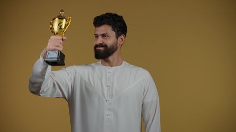 تشجيع الأنديه والمنتخبات، أنشطة ترفيهية ممتعة، النظر إلى الكاميرا بإيماءات الفرح والسعادة لفوز الفريق المفضل، رجل عربي خليجي اماراتي مبتسم يرتدي الزي الكاجوال يقفز عاليا ويمسك بيده الكأس الذهبي، تعابير الانتصار والحماس، خلفية صفراء