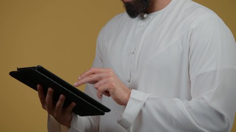 إيماءات الاندماج والتفاعل، استخدام التكنولوجيا الحديثة والمتطورة،  تصوير لرجل عربي خليجي اماراتي يرتدي الكندورة يمسك بيده التابلت المحمول، دمج التقنيات الحديثه فى الحياة اليومية ،خلفية صفراء