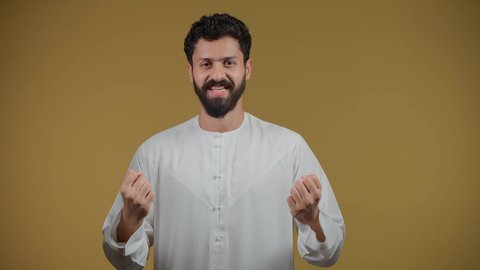 النظر إلى الكاميرا بإيماءات الفرح والسعادة لفوز الفريق المفضل،  أنشطة ترفيهية ممتعة، تشجيع الأنديه والمنتخبات، رجل عربي خليجي اماراتي مبتسم يرتدي الكندورة يرفع قبضة اليد بتعابير الانتصار والحماس، خلفية صفراء