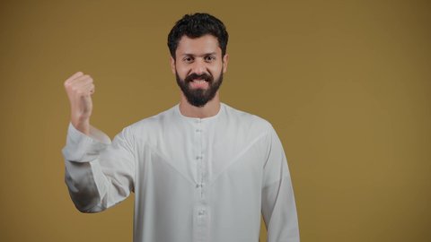 Fun recreational activities, looking at the camera with gestures of joy and happiness for the victory of the favorite team, cheering for clubs and national teams, a smiling Arab Gulf Emirati man wearing a kandura raising his fist with expressions of victory and enthusiasm, yellow background