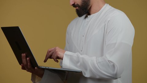 Browsing social media, watching movies and TV series, a picture of a young Emirati Gulf Arab man wearing a kandura holding a laptop, the concept of hard work and completing required tasks, making video calls via laptop, yellow background