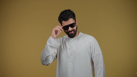 الأناقة والوسامة والاهتمام بالمظهر الخارجي، تصوير لرجل عربي خليجي اماراتي يرتدي الكندورة يقف بإيماءات الثقة بالنفس ويضع نظارة شمسية سوداء، استخدام العدسات الطبية لحماية العين من أشعة الشمس، خلفية صفراء