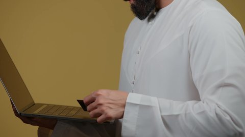 اجراء اجتماع عمل اونلاين، استعمال التكنولوجيا الحديثة والمتطورة، استخدام اللابتوب في إنجاز مهام العمل عن بعد، تصوير لرجل عربي خليجي اماراتي يرتدي الكندورة يستخدم جهاز الحاسوب بإيماءات التركيز، خلفية صفراء