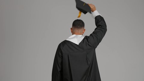 التعليم في الامارات العربية المتحدة، الشعور بالفخر والفرح بالوصول لحلم التخرج, تصوير من الخلف لشاب خريج عربي خليجي اماراتي يرتدي عباءة يرفع قبعة التخرج، الاحتفال بإنهاء المرحلة الجامعية، خلفية رماديه