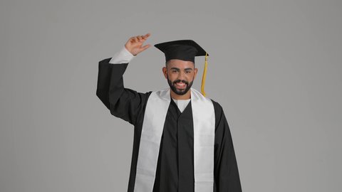 الاحتفال بإنهاء المرحلة الجامعية، الشعور بالفخر والفرح بالوصول لحلم التخرج, التعليم في الامارات العربية المتحدة، تصوير من الخلف لشاب خريج عربي خليجي اماراتي يرتدي عباءة يرفع قبعة التخرج، خلفية رماديه