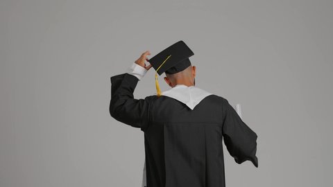 الشعور بالفخر والفرح بالوصول لحلم التخرج, التعليم في الامارات العربية المتحدة، تصوير من الخلف لشاب خريج عربي خليجي اماراتي يرتدي عباءة يرفع شهادة وقبعة التخرج، الاحتفال بإنهاء المرحلة الجامعية، خلفية رماديه