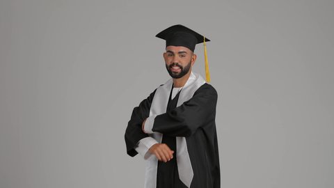 مفهوم التفوق والنجاح والاجتهاد، الاحتفال بإنهاء المرحلة الجامعية، تصوير لشاب خريج عربي خليجي اماراتي مبتسم يرتدي القبعة وعباءة التخرج يقف بأيدي متقاطعة ينظر الى الكاميرا بتعابير الثقة بالنفس، خلفية رمادية