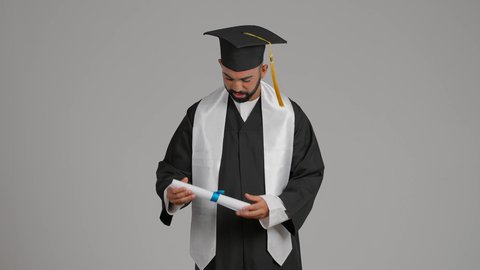 الاحتفال بإنهاء الدراسة الجامعية، الوصول لحلم النجاح، مفهوم الاجتهاد والتفوق، مناسبة النجاح في الجامعة، تعابير السعادة والسرور، جودة التعليم في الامارات، تصوير لشاب خريج عربي خليجي اماراتي مبتسم يرتدي عباءة وقبعة التخرج ينظر الي الشهاده بايماءات الفخر ، خلفية رماديه