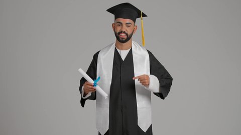 الوصول لحلم النجاح، مفهوم الاجتهاد والتفوق، مناسبة النجاح في الجامعة، تعابير السعادة والسرور، الاحتفال بإنهاء الدراسة الجامعية، جودة التعليم في الامارات، تصوير لشاب خريج عربي خليجي اماراتي مبتسم يرتدي عباءة وقبعة التخرج يشير الي الشهاده بايماءات الفخر ، خلفية رماديه