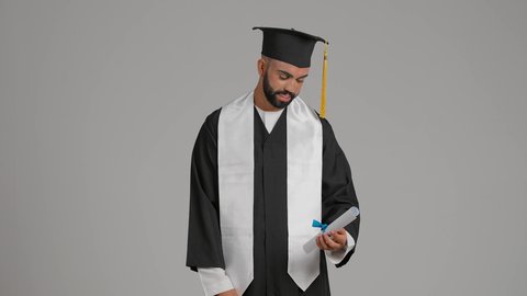 جودة التعليم في الامارات، الوصول لحلم النجاح، تعابير السعادة والسرور، الاحتفال بإنهاء الدراسة الجامعية،  مفهوم الاجتهاد والتفوق،مناسبة النجاح في الجامعة، تصوير لشاب خريج عربي خليجي اماراتي مبتسم يرتدي عباءة وقبعة التخرج يمسك الشهاده بيده ، يرفع اصبع السبابه بايماءات الفخر ، خلفية رماديه