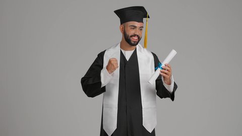 مفهوم الاجتهاد والتفوق، جودة التعليم في الامارات، الوصول لحلم النجاح، تعابير السعادة والسرور، الاحتفال بإنهاء الدراسة الجامعية، مناسبة النجاح في الجامعة، تصوير لشاب خريج عربي خليجي اماراتي مبتسم يرتدي عباءة وقبعة التخرج يمسك الشهاده بيده ، يرفع قبضه يده بايماءات الفخر ، خلفية رماديه