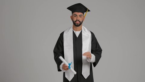 جودة التعليم في الامارات، الوصول لحلم النجاح، تعابير السعادة والسرور، مناسبة النجاح في الجامعة، الاحتفال بإنهاء الدراسة الجامعية، مفهوم الاجتهاد والتفوق، تصوير لشاب خريج عربي خليجي اماراتي مبتسم يرتدي عباءة وقبعة التخرج يمسك بيده الشهادة بايماءات الجودة والامتياز ، خلفية رماديه