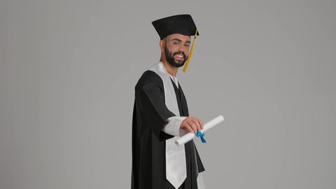 Quality of education in the UAE, achieving the dream of success, expressions of happiness and joy, the occasion of university success, celebrating the completion of university studies, the concept of diligence and excellence, a photograph of a smiling young Emirati Gulf Arab graduate wearing a graduation gown and cap, holding a diploma, gray background.