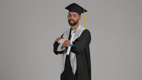 مفهوم التفوق والنجاح والاجتهاد، الاحتفال بإنهاء المرحلة الجامعية، تصوير لشاب خريج عربي خليجي اماراتي مبتسم يرتدي القبعة وعباءة التخرج يقف بأيدي متقاطعة ينظر الى الكاميرا بتعابير الثقة بالنفس، يمسك بيده الشهاده، خلفية رمادية