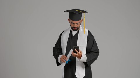 استخدام التقنية الحديثة، الاحتفال بإنهاء الدراسة الجامعية، الاحتفاظ بالذكريات السعيدة، تصوير لشاب خريج عربي خليجي اماراتي يرتدي عباءه وقبعة التخرج يقوم بالتقاط صورة سيلفي تذكارية عبر الهاتف المحمول، خلفية رماديه