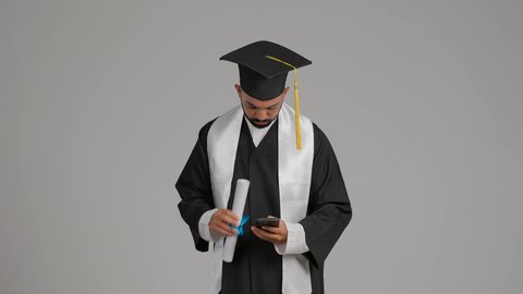الاحتفاظ بالذكريات السعيدة، استخدام التقنية الحديثة، الاحتفال بإنهاء الدراسة الجامعية، تصوير لشاب خريج عربي خليجي اماراتي يرتدي عباءه وقبعة التخرج يقوم بالتقاط صورة سيلفي تذكارية عبر الهاتف المحمول، خلفية رماديه