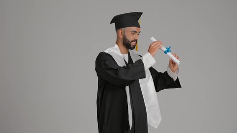 الفخر والفرح بالوصول لحلم التخرج, التطلع إلى المستقبل بعد التخرج، تصوير لشاب خريج عربي سعودي خليجي يرتدي ثياب التخرج ينظر من خلال شهادة التفوق، الاحتفال بانتهاء المرحلة الجامعية، خلفية رماديه